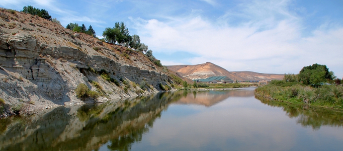 [View from a bridge across the river. On the left bank the rocky land is approximately 60 feet high while on the right the grassy land is just a few feet above the water. The river bends to the right in the distance and behind it are sandstone colored mountains.]